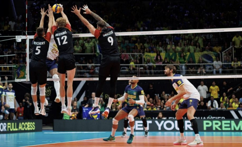 Volleyballspiel: drei Spieler springen zum Block am Netz, Gegner wartet auf Annahme, Zuschauer.